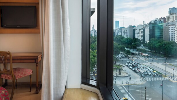A hotel room with a large window offering a city view, a flat-screen TV mounted on the wall, and a wooden chair with a patterned cushion. - Imperial Park Hotel (Buenos Aires)