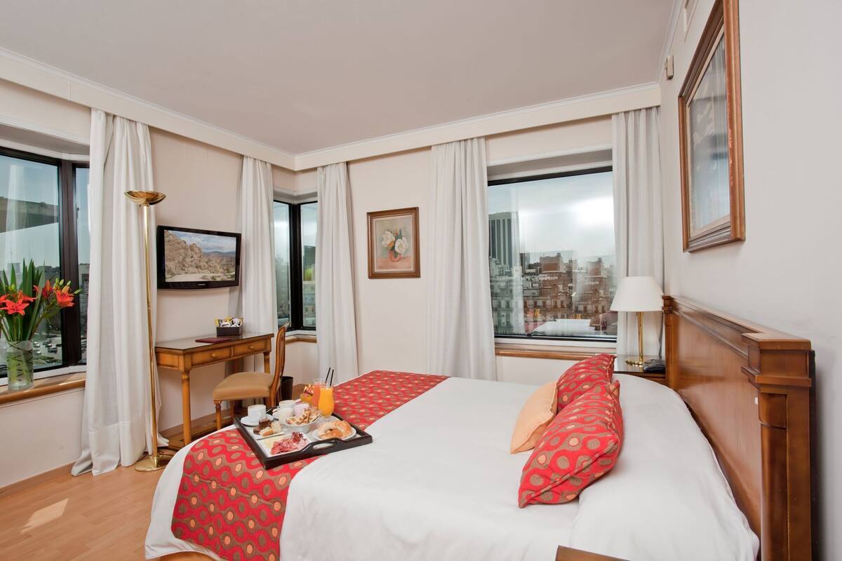 Quarto de hotel com cama, escrivaninha, cadeira, janela com vista para a cidade e bandeja com itens de café da manhã.