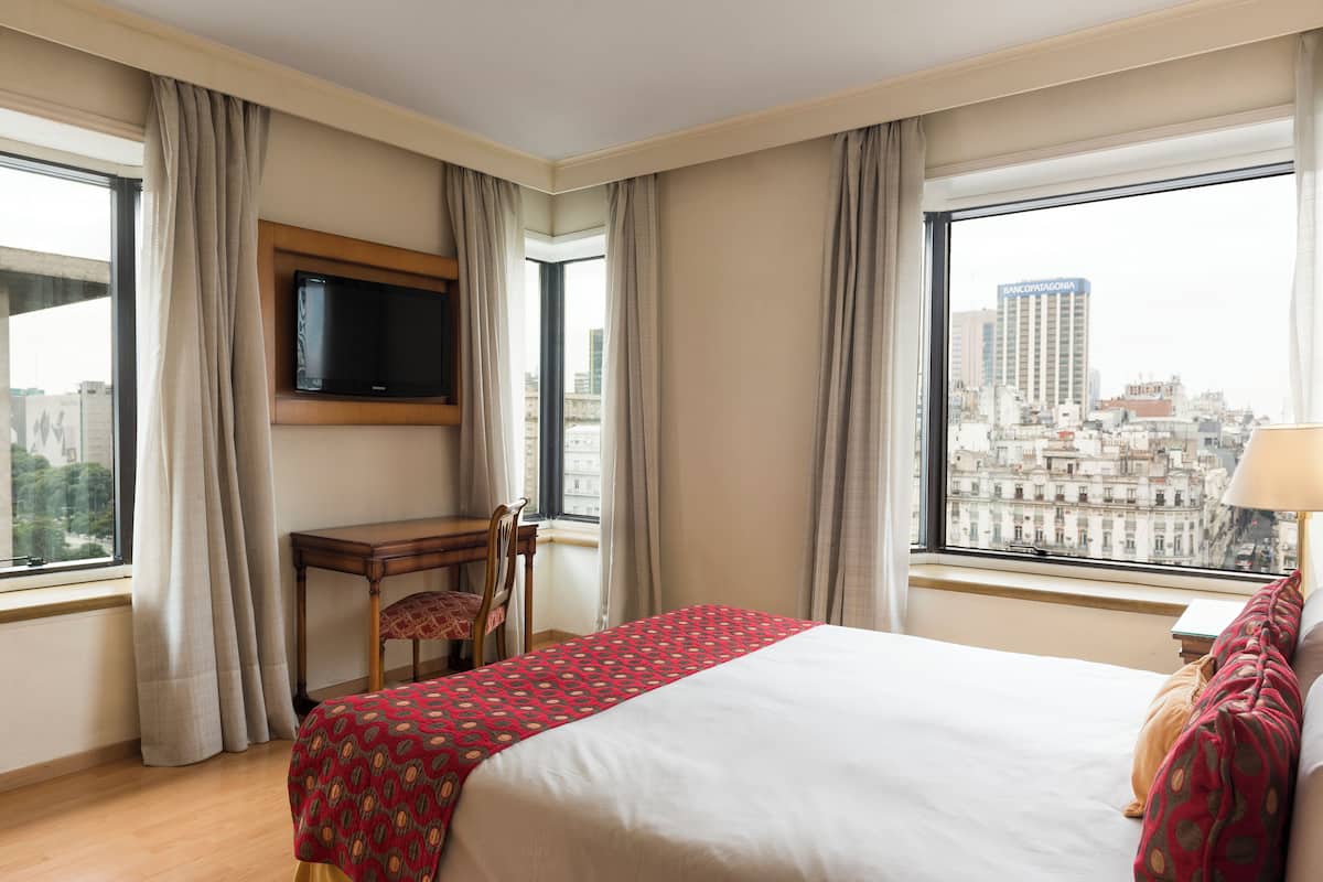 Quarto de hotel com cama, escrivaninha, cadeira, televisão e uma grande janela com vista para a cidade.