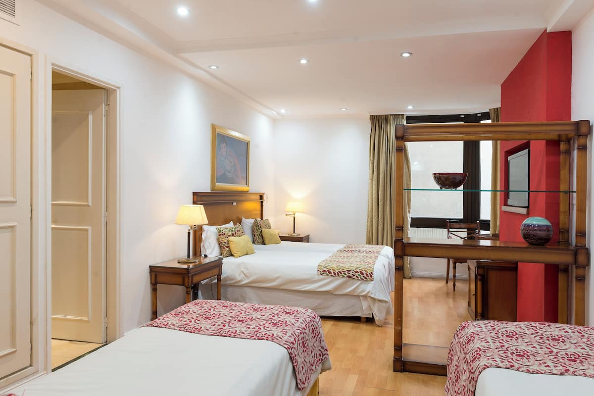 Quarto de hotel com duas camas, uma escrivaninha com cadeira, uma janela com cortinas e uma parede de destaque vermelha.