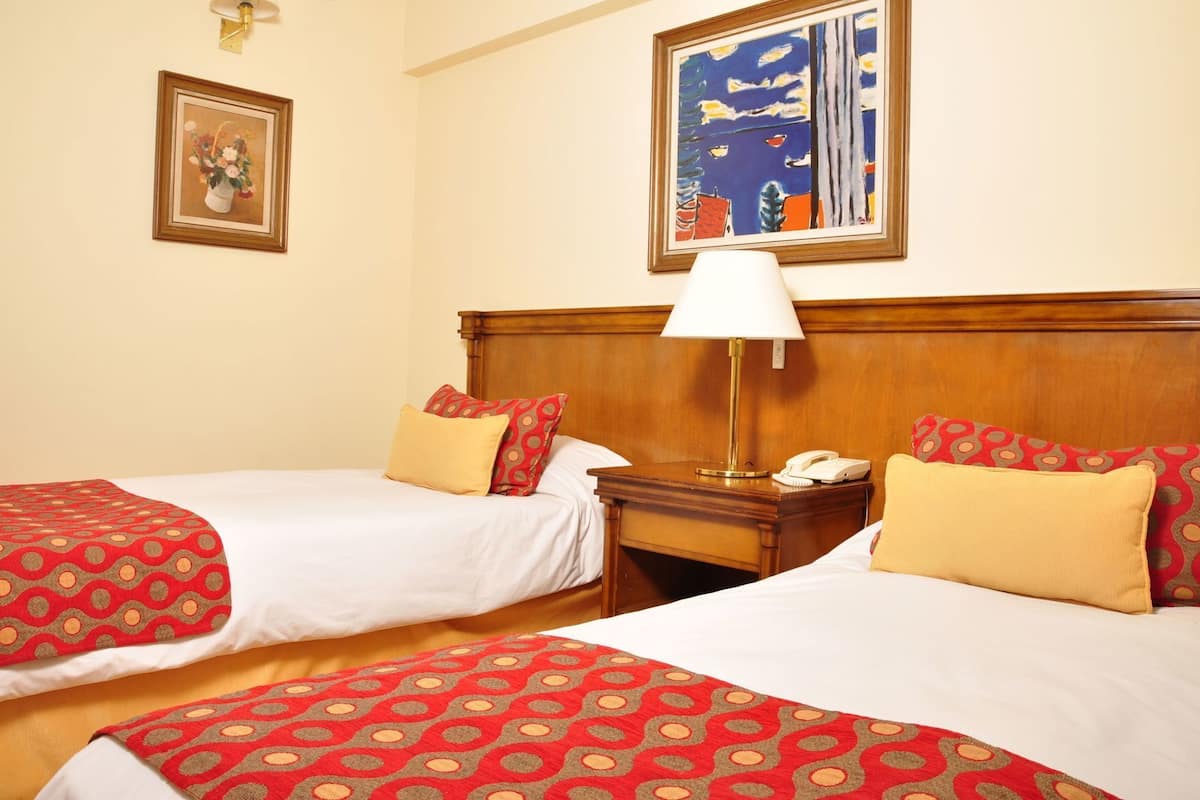 Quarto de hotel com duas camas, uma mesa de cabeceira com abajur, telefone e duas pinturas na parede.