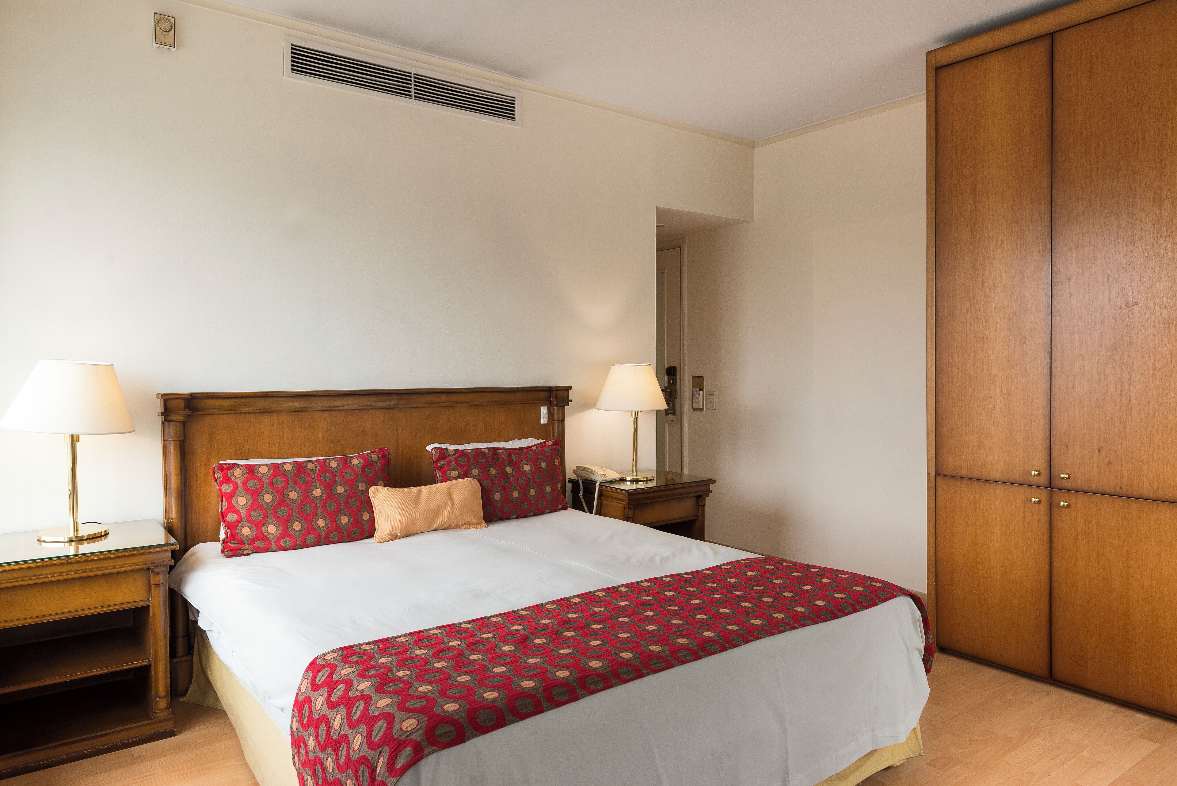 A neatly made bed with a patterned bedspread, two bedside lamps, and a wooden wardrobe.
