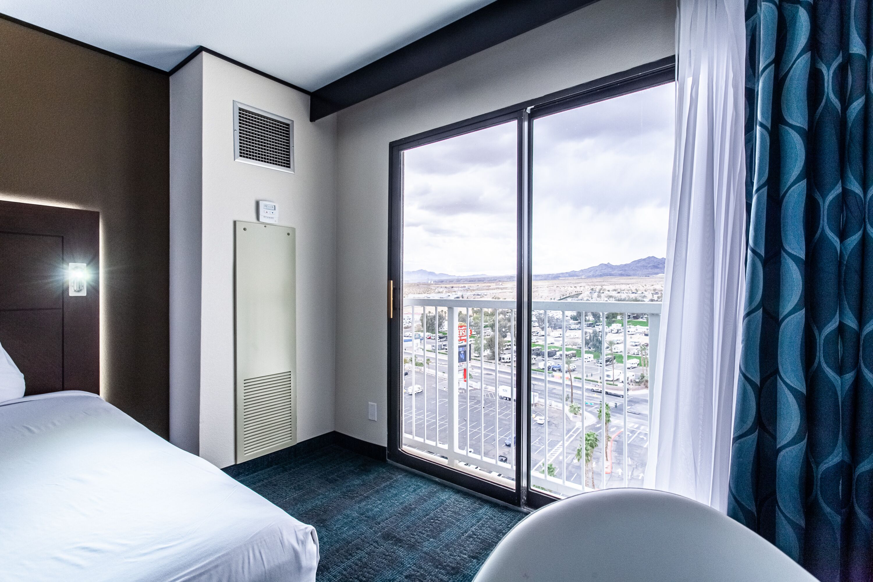 Une chambre d’hôtel avec un lit, une chaise, une grande fenêtre donnant sur un paysage urbain et une grille d’aération au mur.