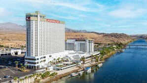 Un hotel de gran altura con un llamativo letrero del Casino Riverside, situado junto a un río con un puente y colinas desérticas al fondo.