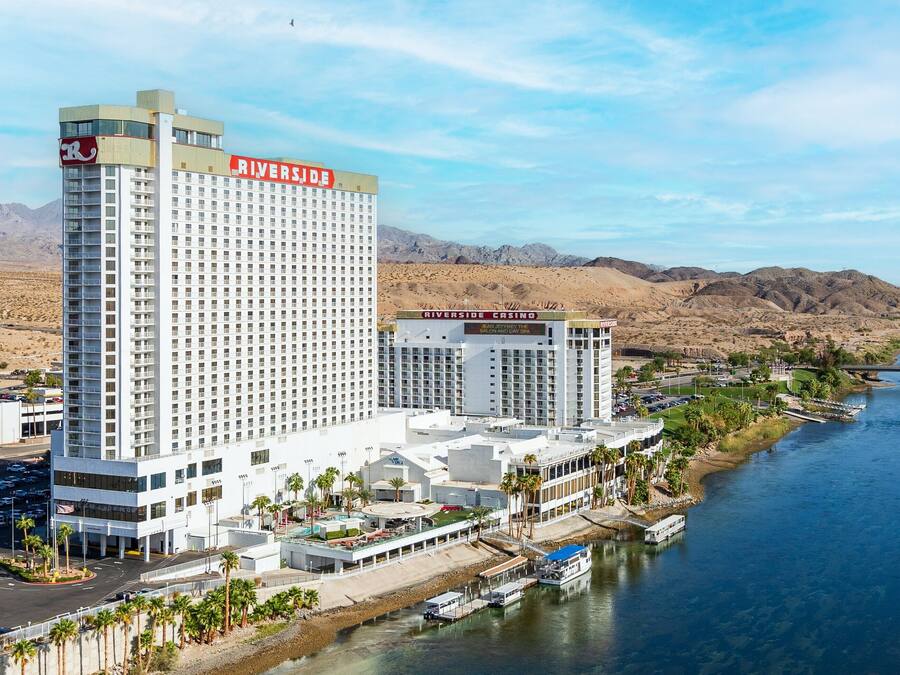 A high-rise hotel with a prominent Riverside Casino sign, located beside a river with a bridge and desert hills in the background.