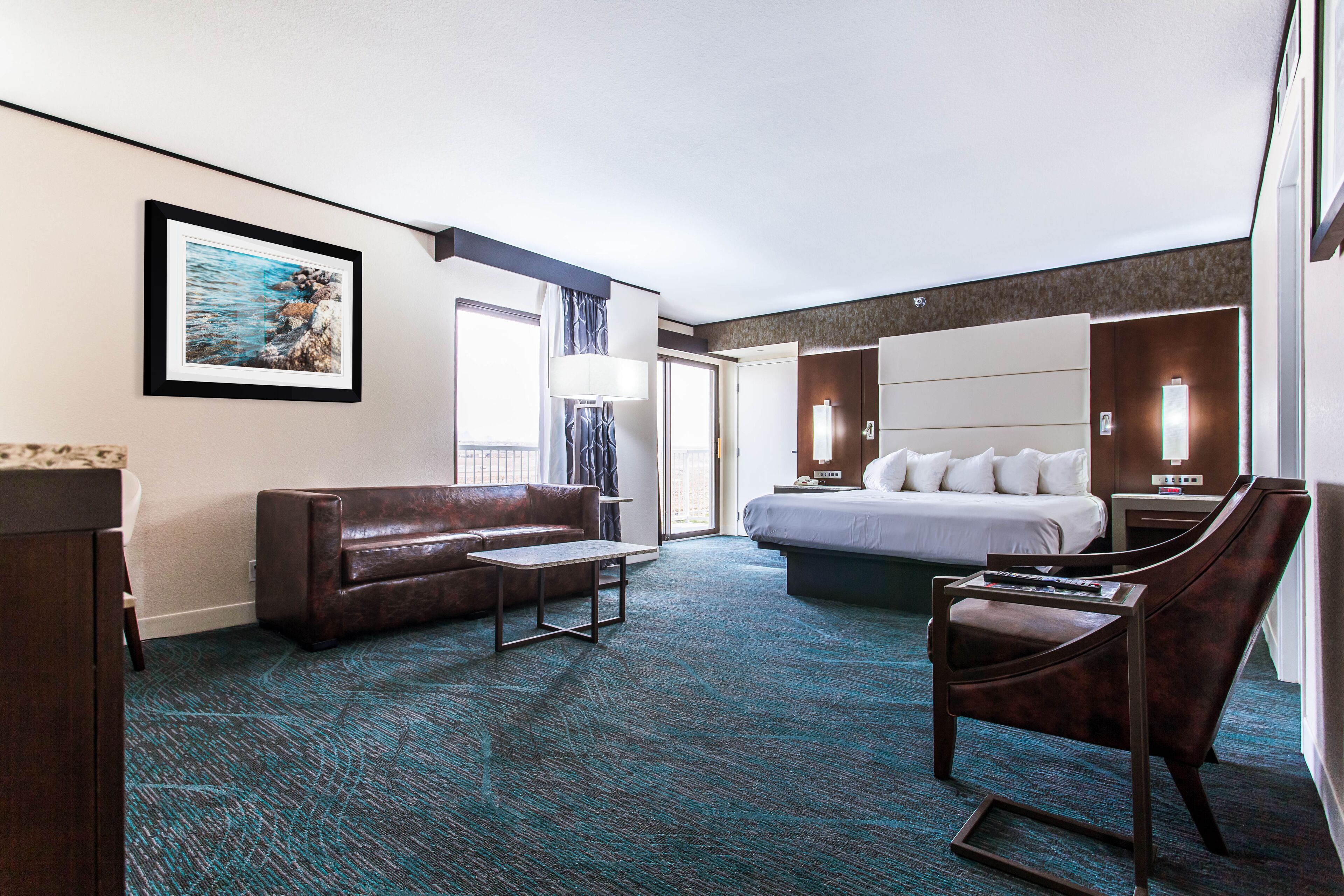 A hotel room with a bed, a sofa, a desk, and a chair. There is a painting on the wall and a window with curtains.