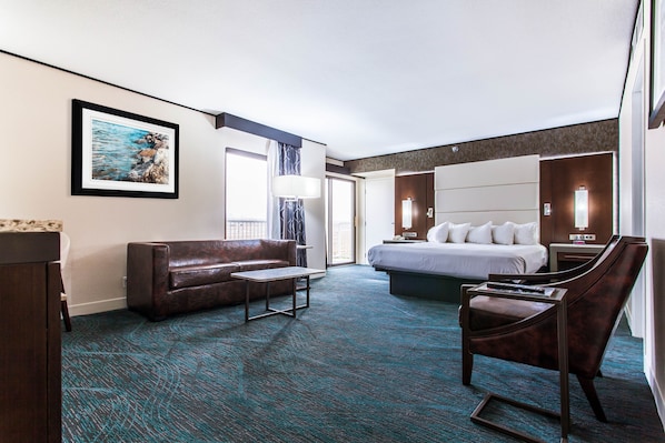 Habitación de hotel con cama, sofá, escritorio y una silla. Se observa un cuadro en la pared y una ventana con cortinas.