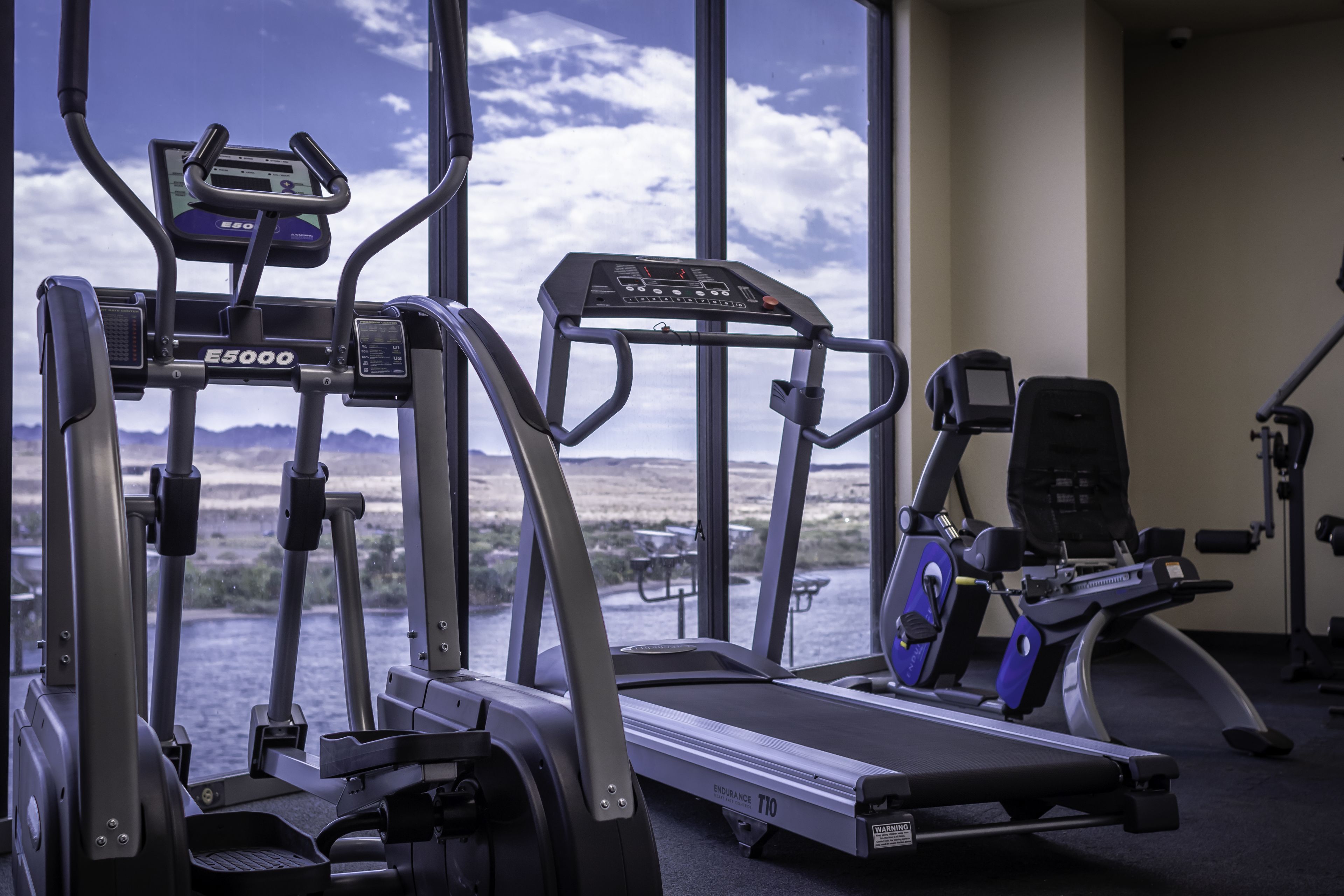 Uma academia com diversos equipamentos de exercício, incluindo esteiras e uma máquina elíptica, com vista para uma paisagem montanhosa através de grandes janelas.