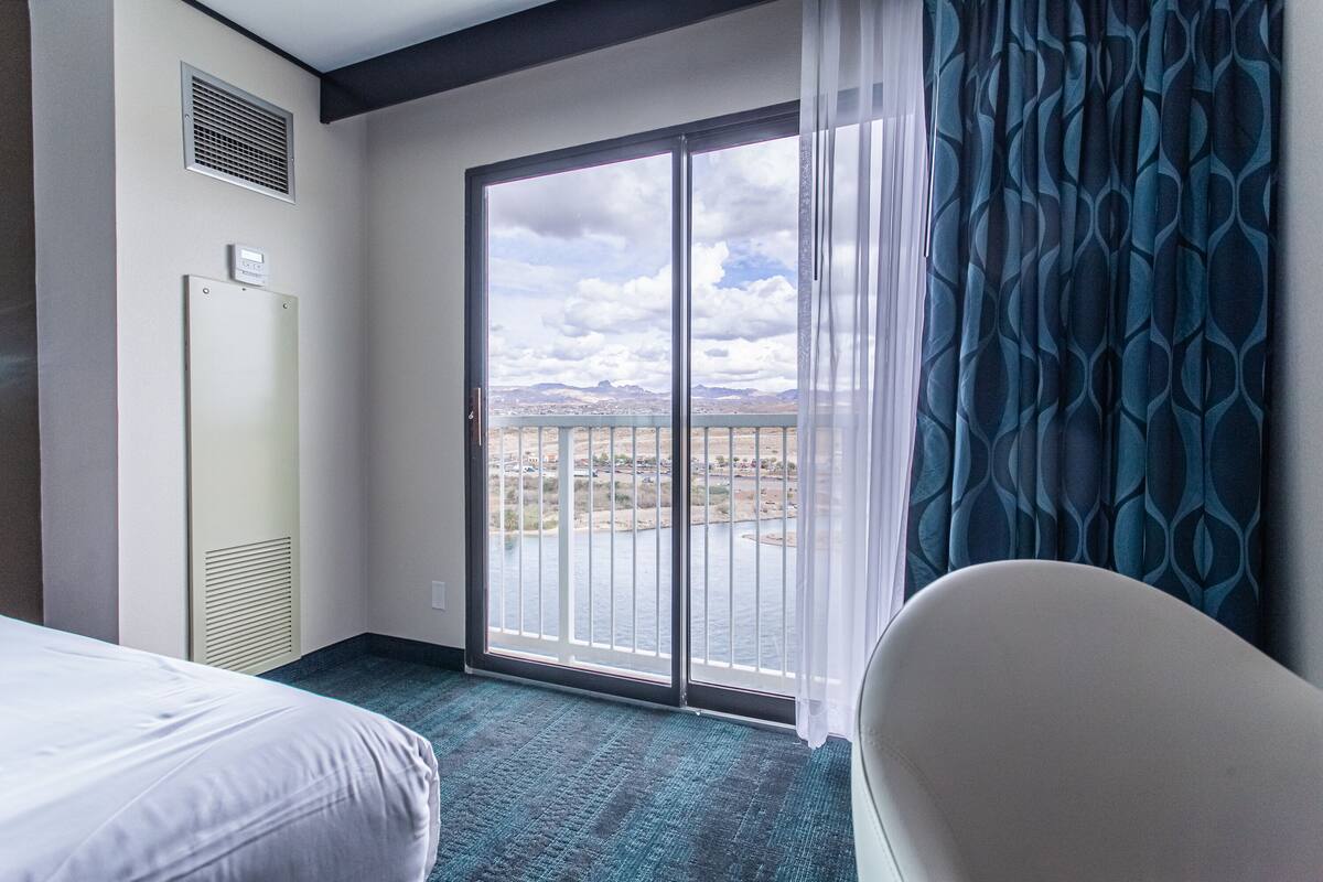A hotel room with a bed, a chair, a large window with a view of a cityscape, and a door with a vent.