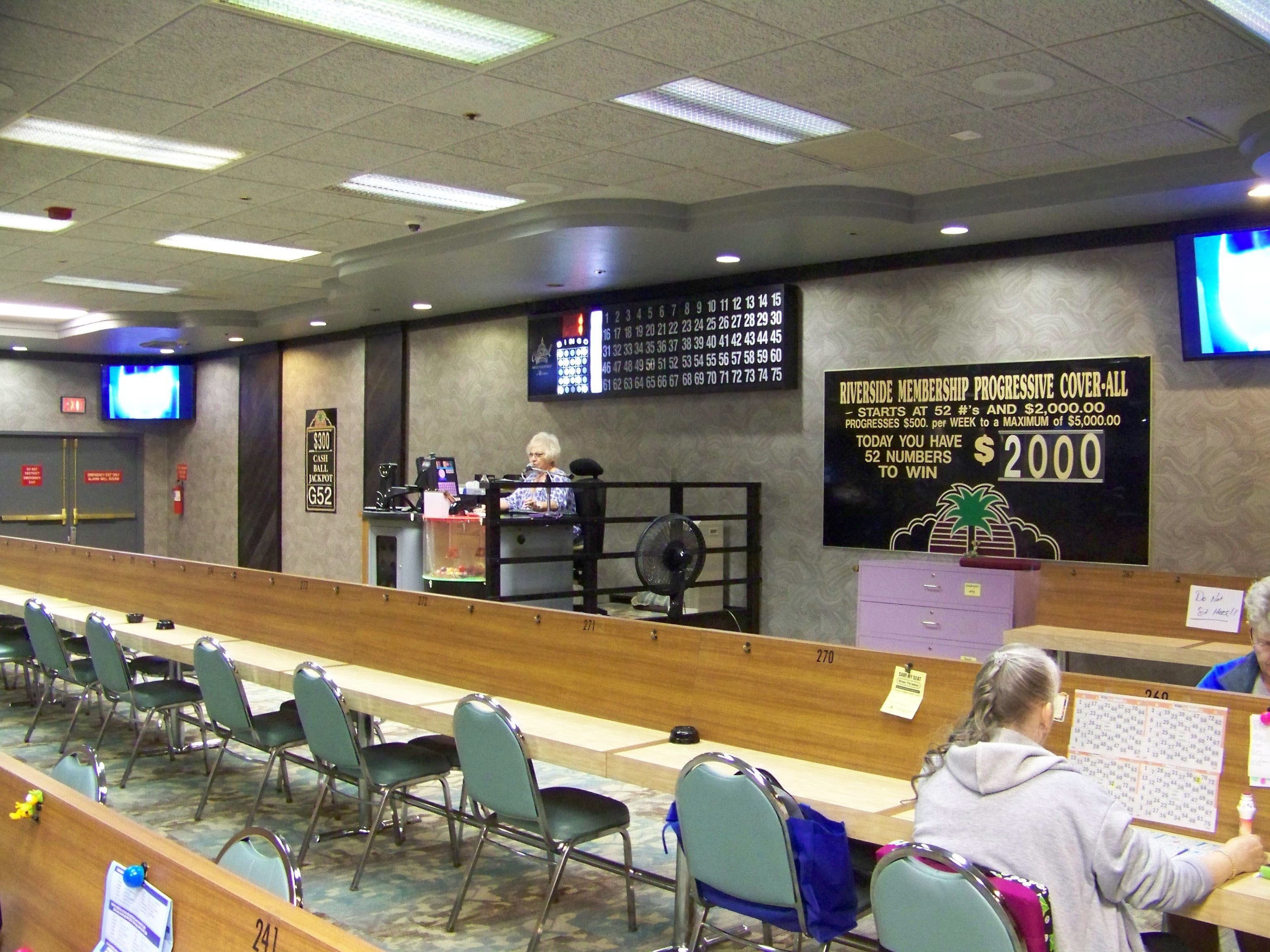 A room with rows of chairs, a counter with a computer, and a large sign about Riverside Membership Progressive Cover-All.