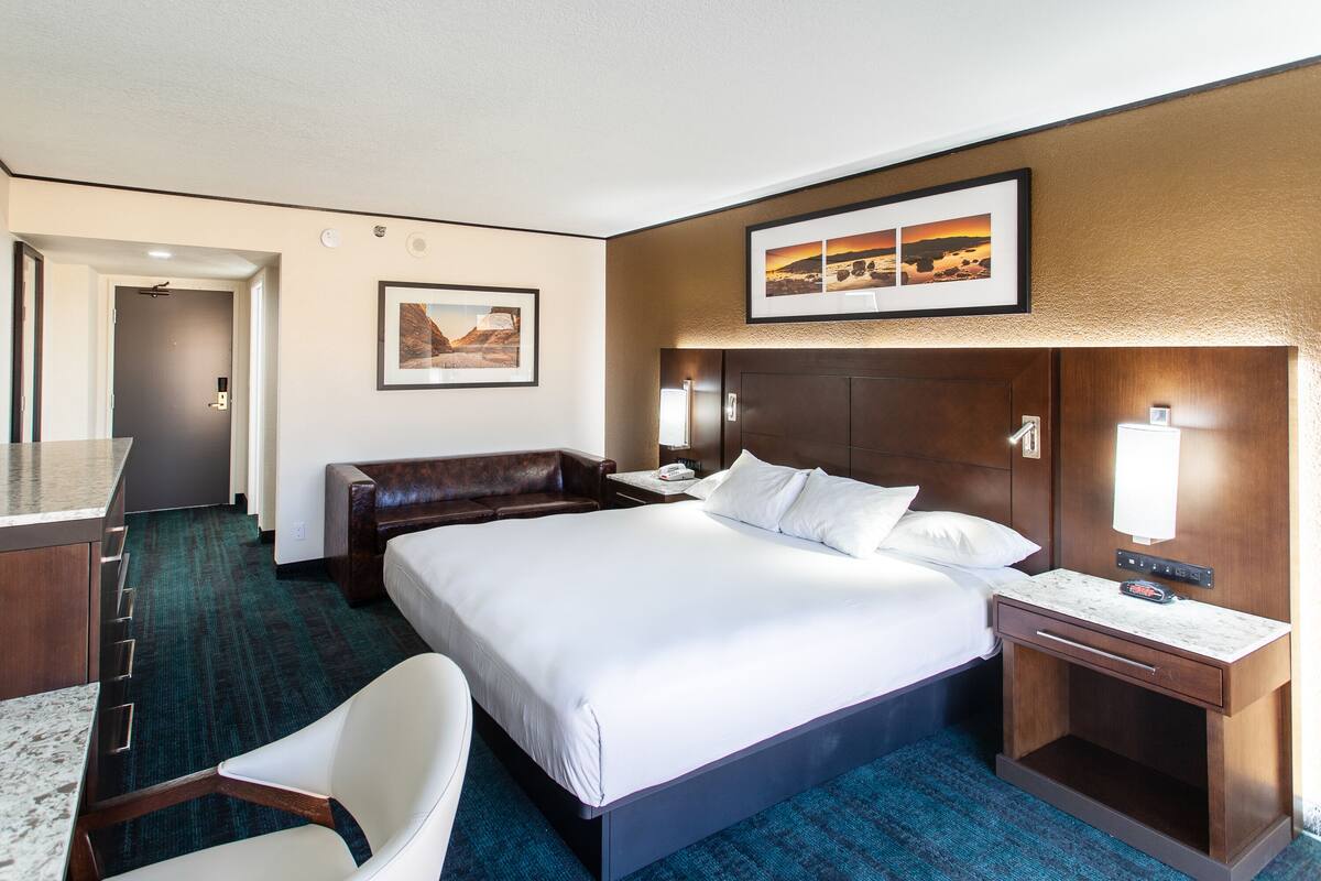 A hotel room with a large bed, a desk, a chair, and a nightstand with a lamp.