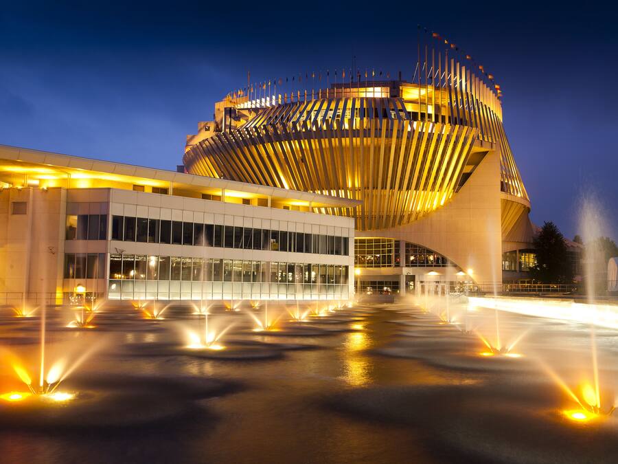 A modern building with a unique curved roof and a series of fountains in front.