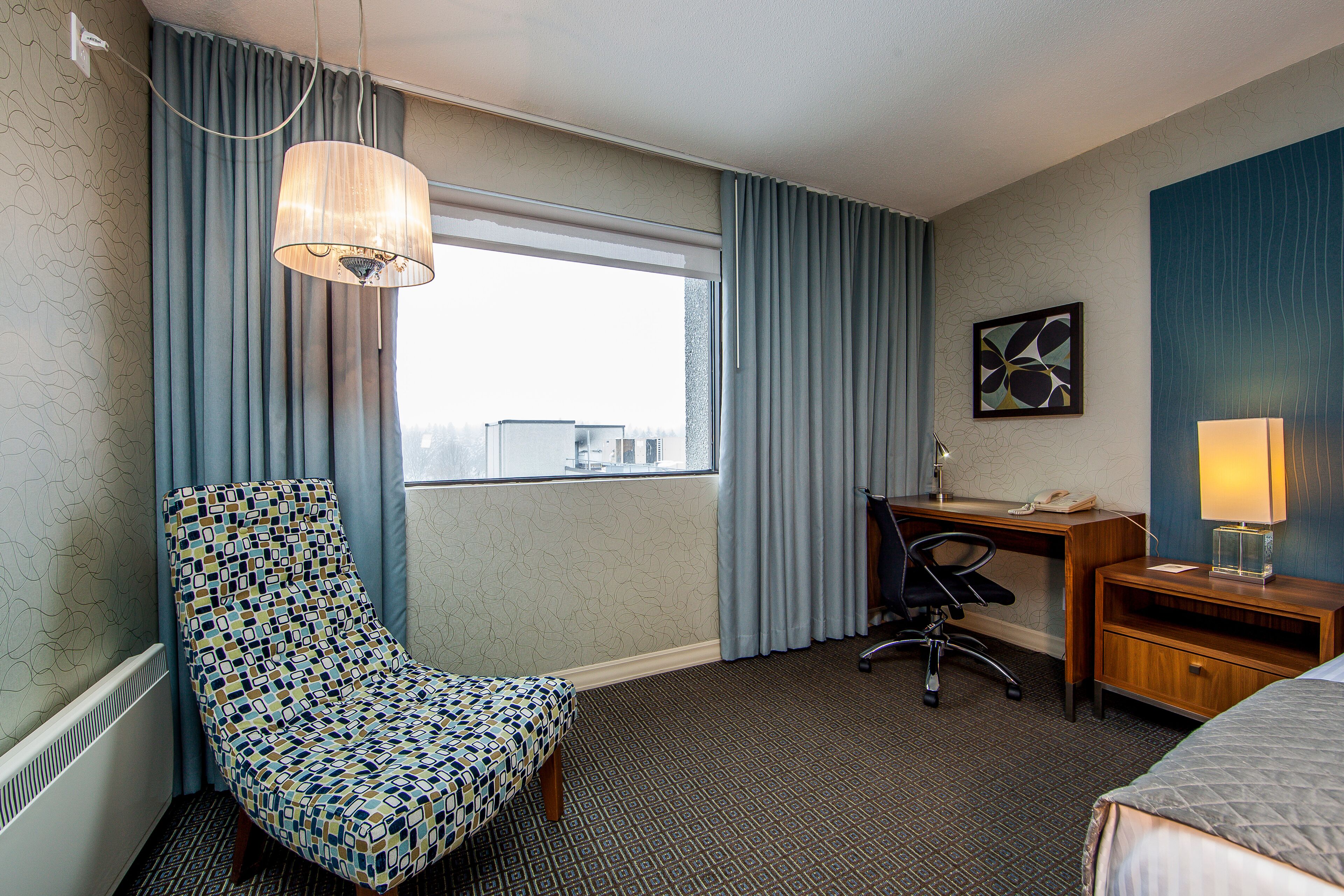 Hotellihuone, jossa on kuviollinen nojatuoli, työpöytä lampun kanssa, sänky ja ikkunasta avautuva näkymä kaupunkiin.