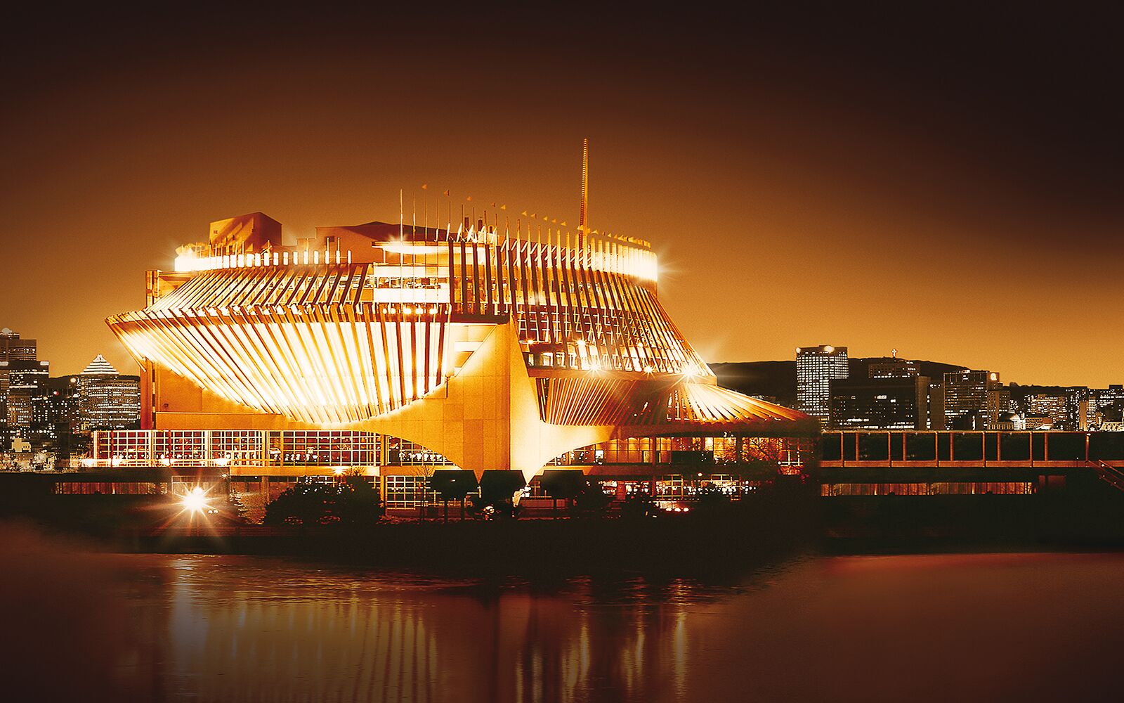 Un bâtiment moderne doté d’une structure de toit originale, entouré d’eau et avec la silhouette de la ville en toile de fond.