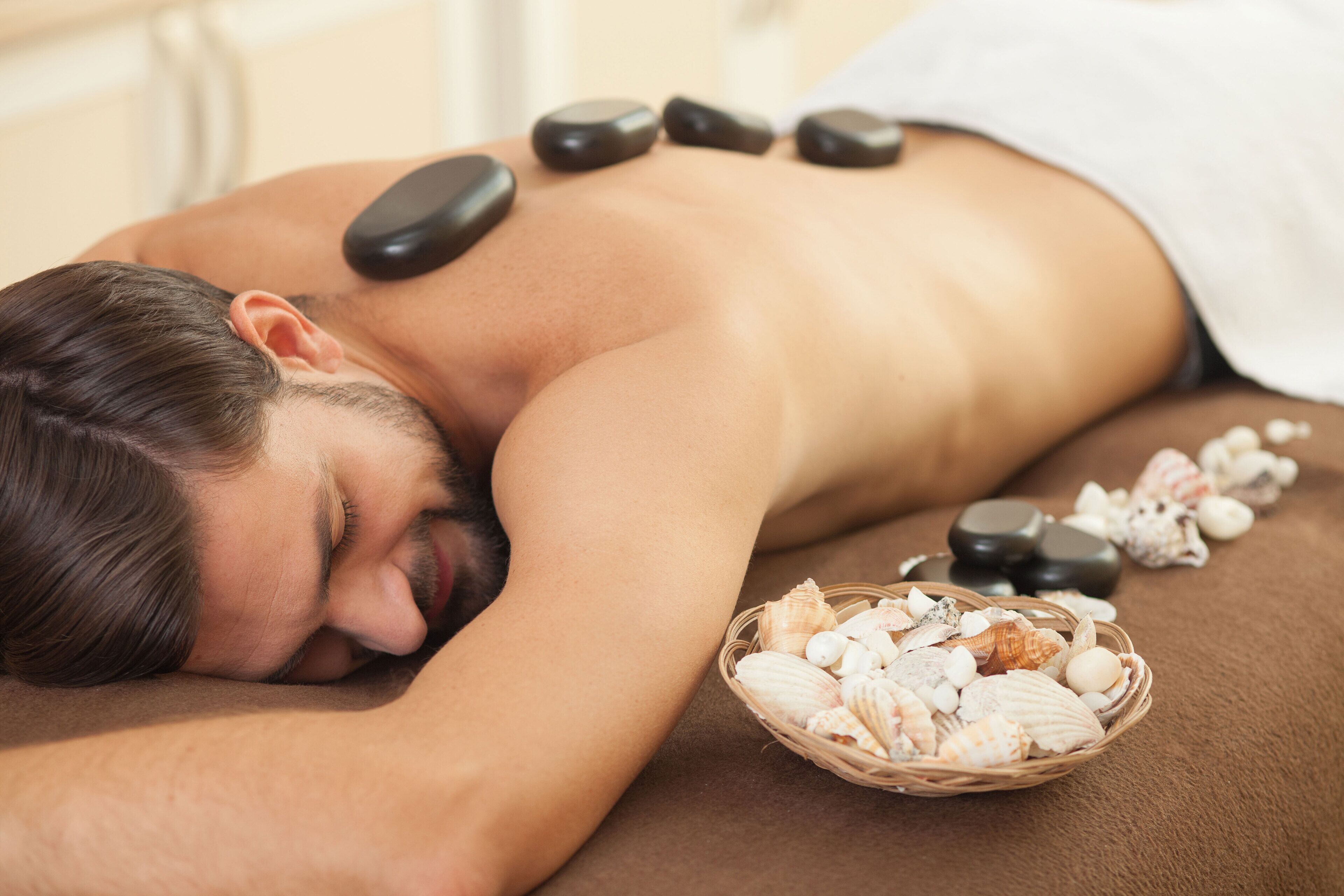 Una persona recibe un masaje con piedras, con una cesta de piedras y conchas cerca.