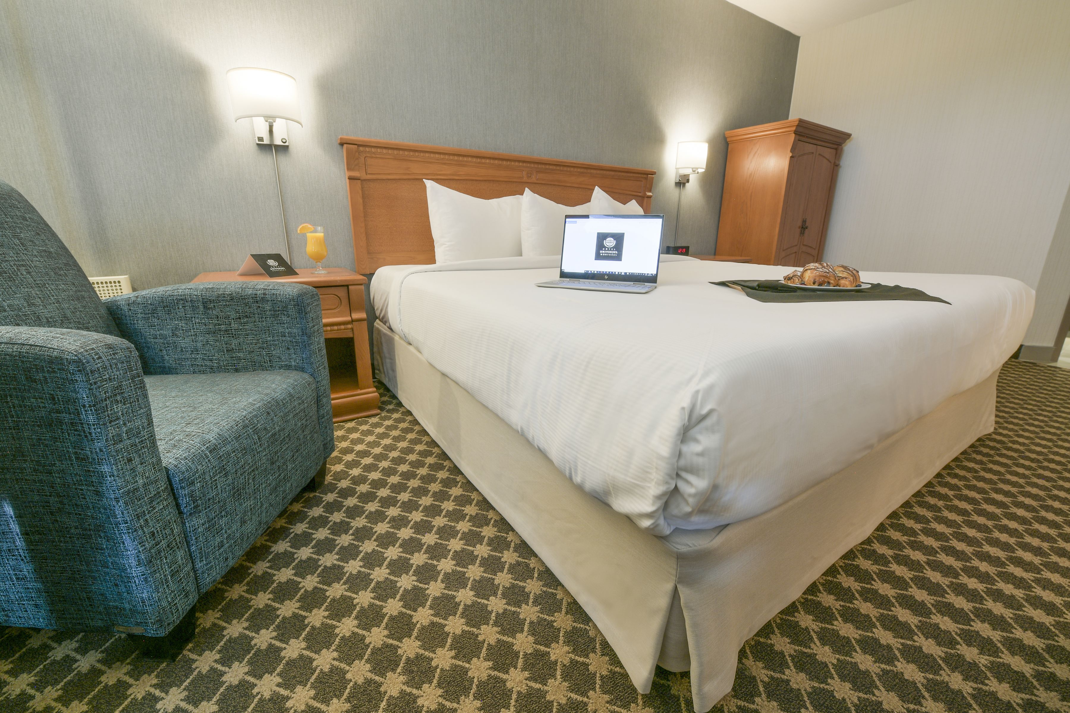 A hotel room with a large bed, a chair, a desk with a laptop, and a nightstand with a lamp.