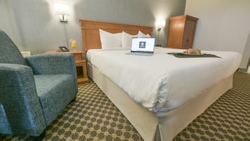 A hotel room with a large bed, a chair, a desk with a laptop, and a nightstand with a lamp.
