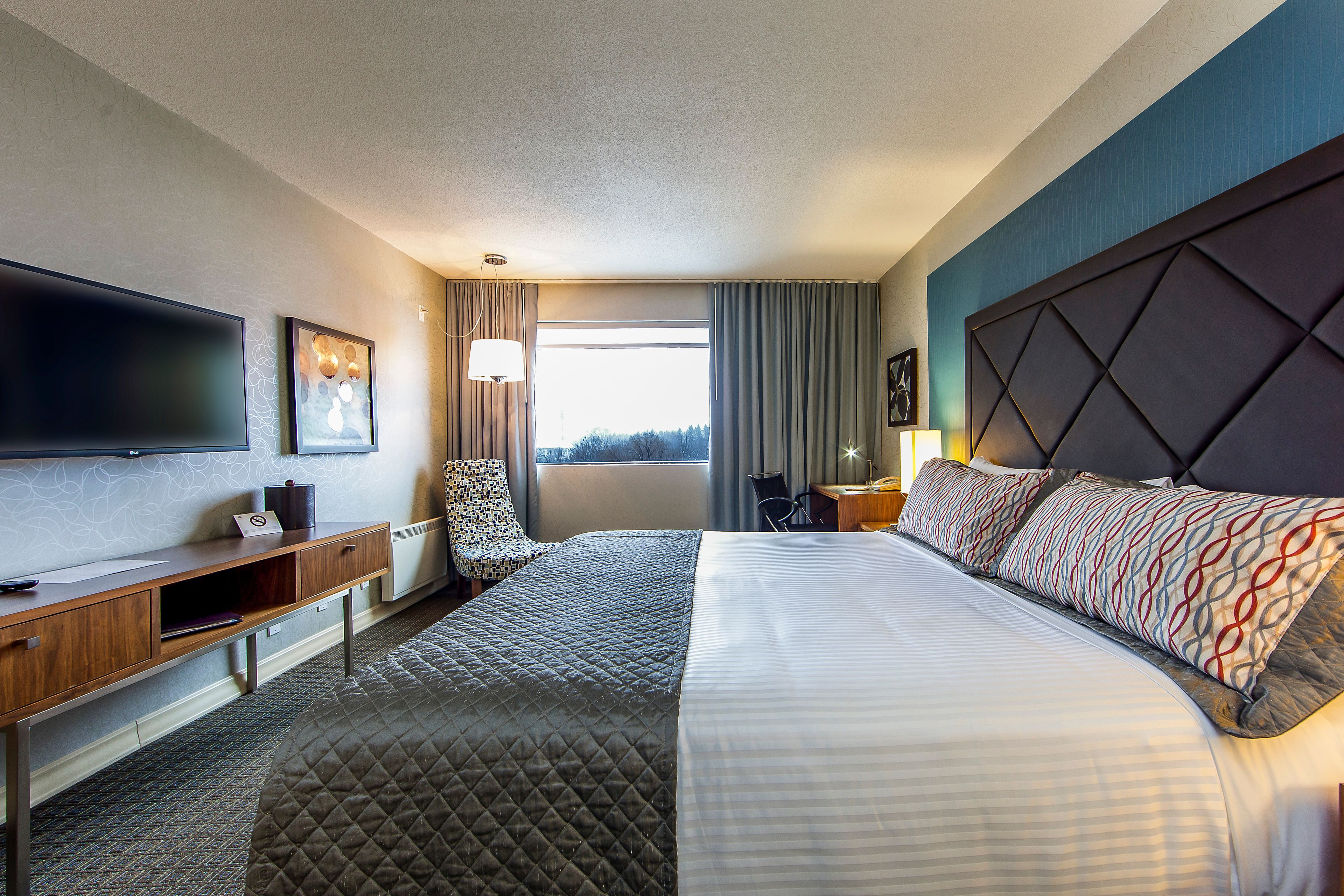 A hotel room with a large bed, a desk with a flat-screen TV, a chair, and a window with curtains.