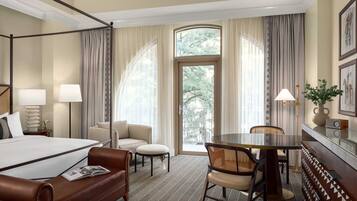 A hotel room with a four-poster bed, a sofa, a dining table with chairs, and a large window with curtains.
