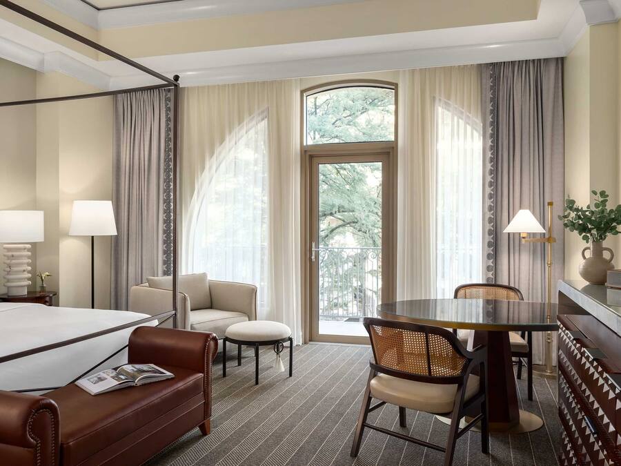 A hotel room with a four-poster bed, a sofa, a dining table with chairs, and a large window with curtains.
