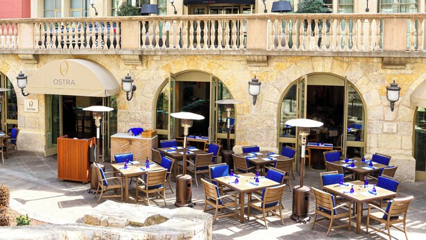Un área de café al aire libre con mesas y sillas, un letrero que dice OSTRA, y balcones con barandillas.