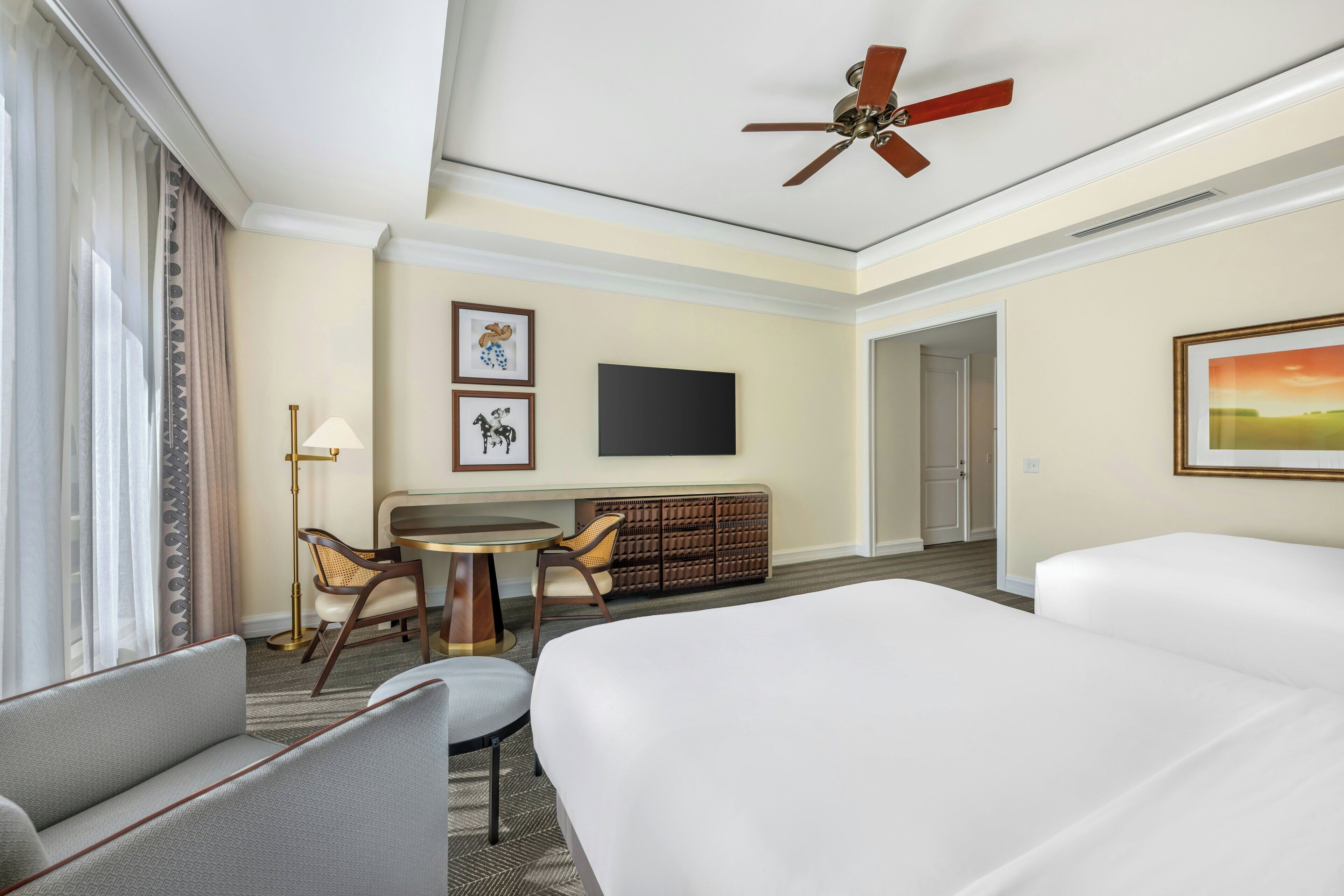 A hotel room with a bed, a desk, two chairs, a television, and a ceiling fan.