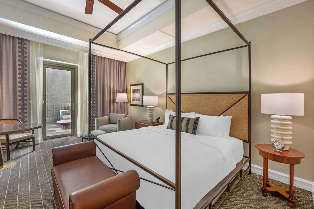 A four-poster bed with a wooden headboard, a small round table with a clock, a sofa, and a desk with a chair in a hotel room.