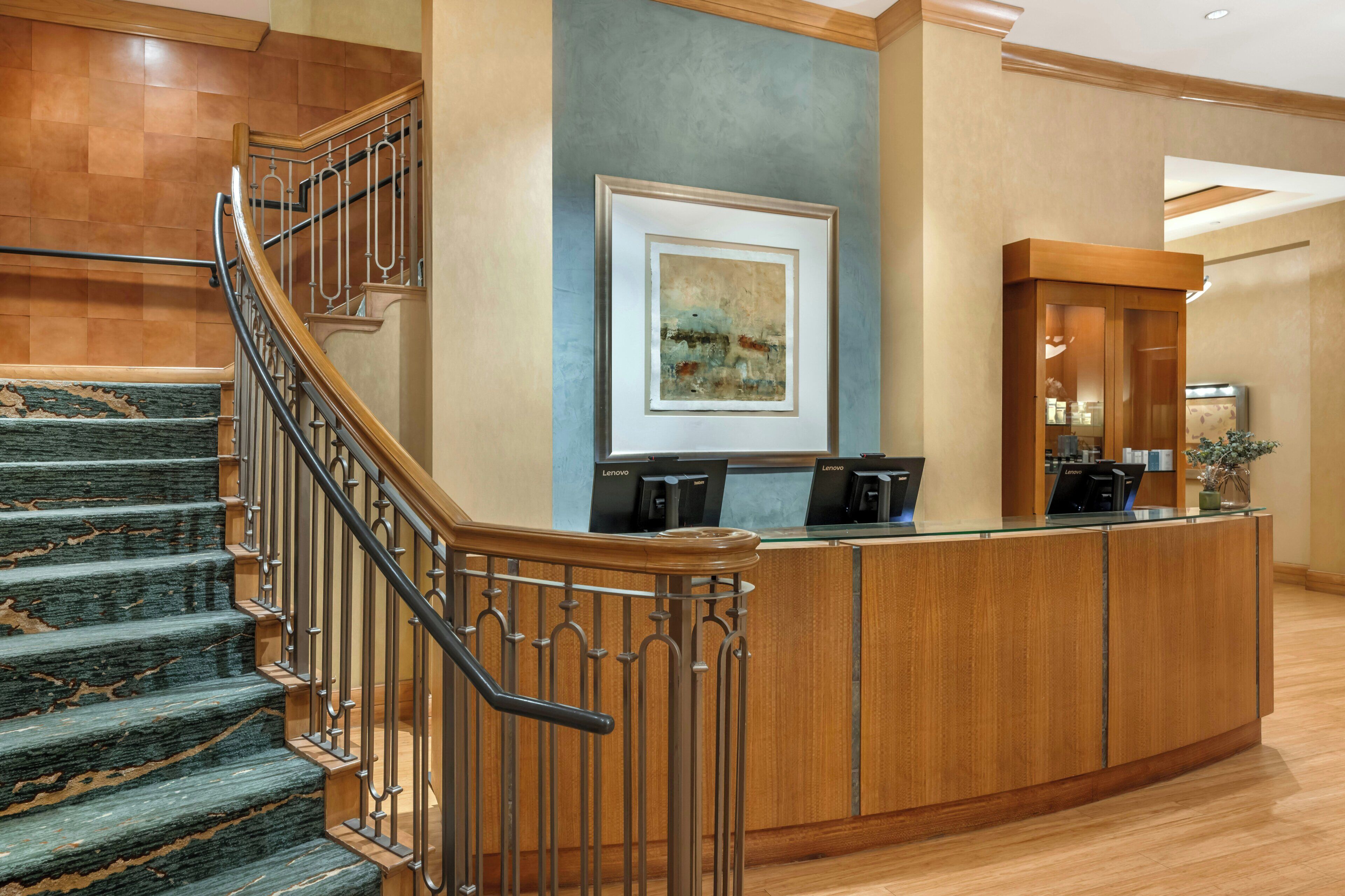 A hotel lobby with a curved reception desk, a staircase with wooden railings, and a framed artwork on the wall.