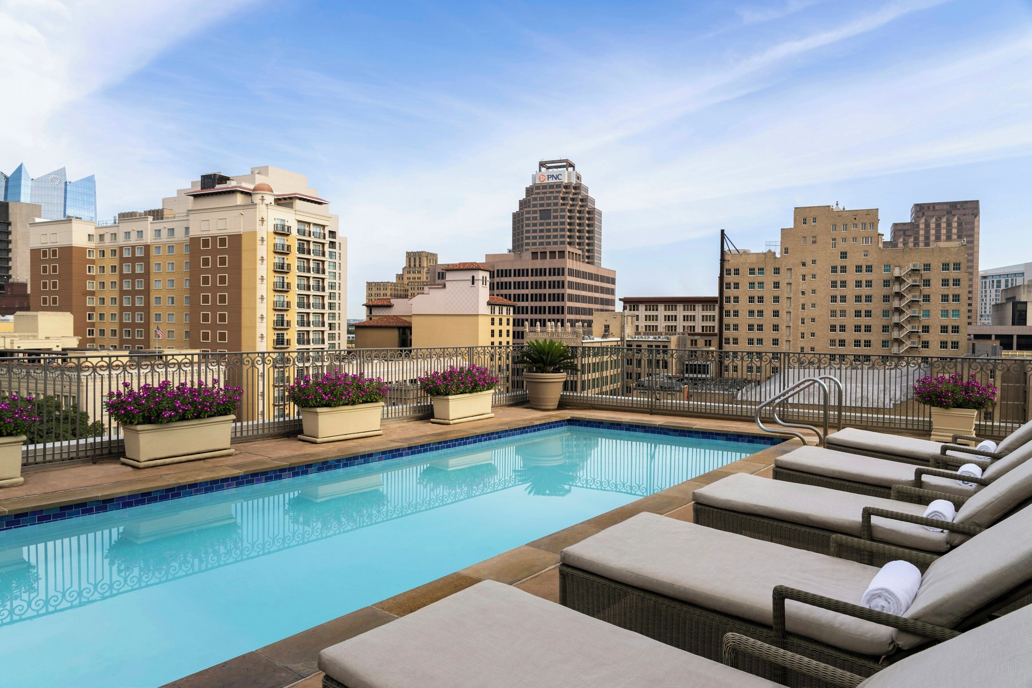 A rooftop pool with lounge chairs, potted plants, and a cityscape view.