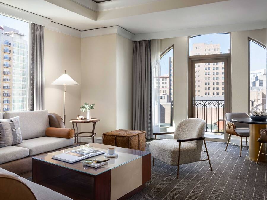 A modern hotel room with a sofa, coffee table, and chairs. It features large windows with city views and a balcony.