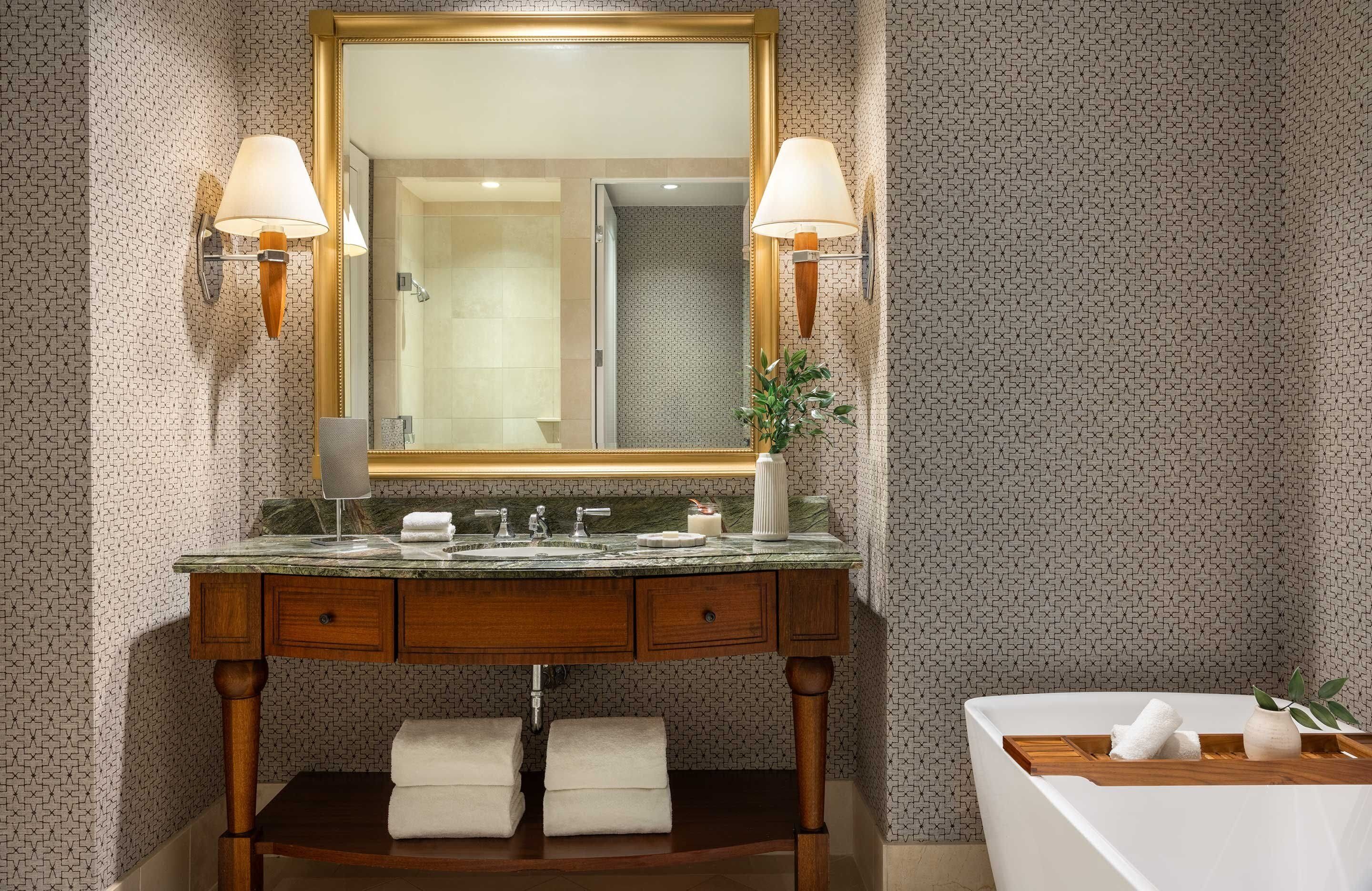 A bathroom with a large mirror, two wall-mounted lamps, a marble countertop, wooden vanity, and a bathtub with a tray.