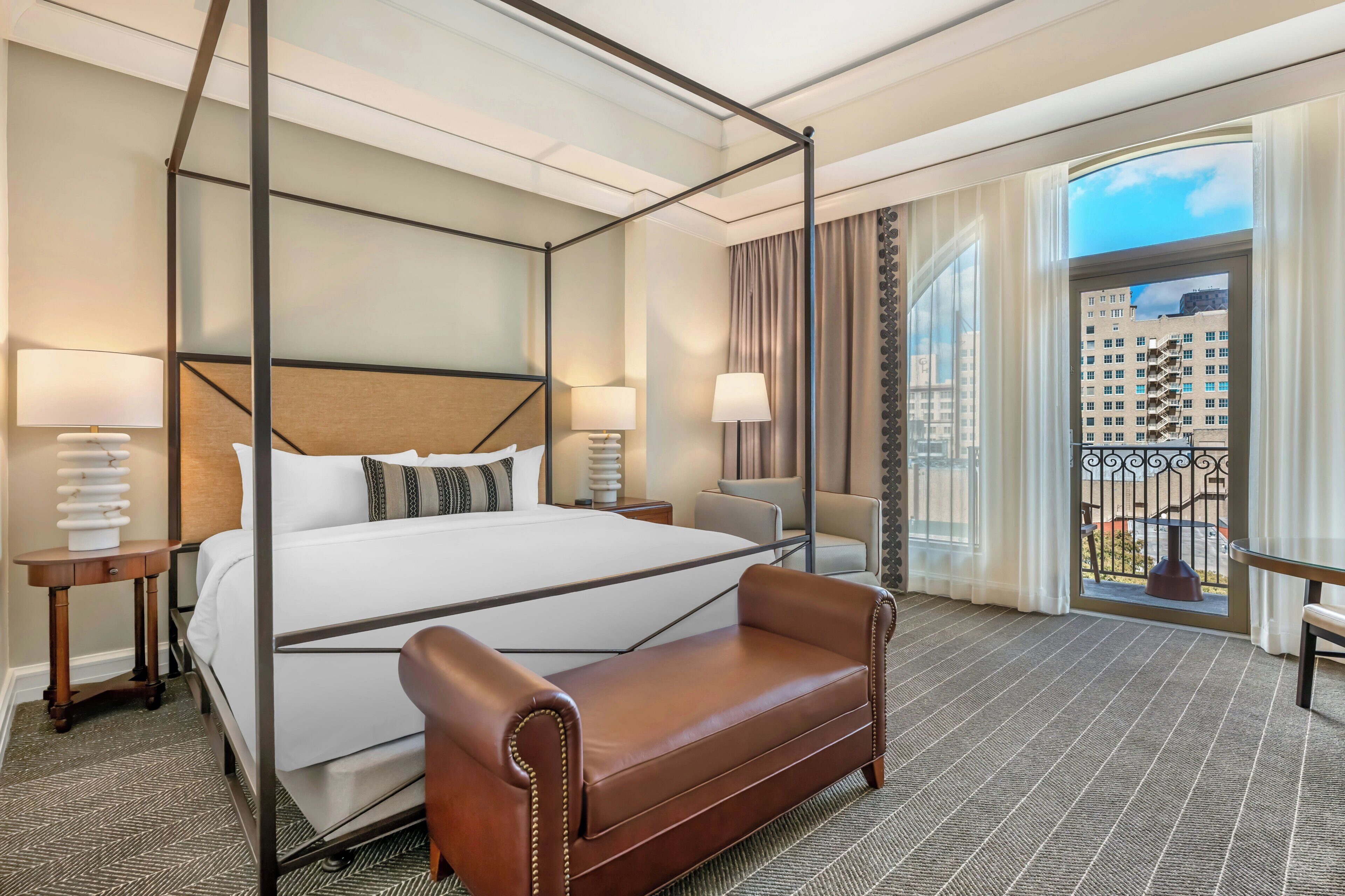 A hotel room with a four-poster bed, a leather sofa, a balcony with a view of buildings, and two bedside lamps.