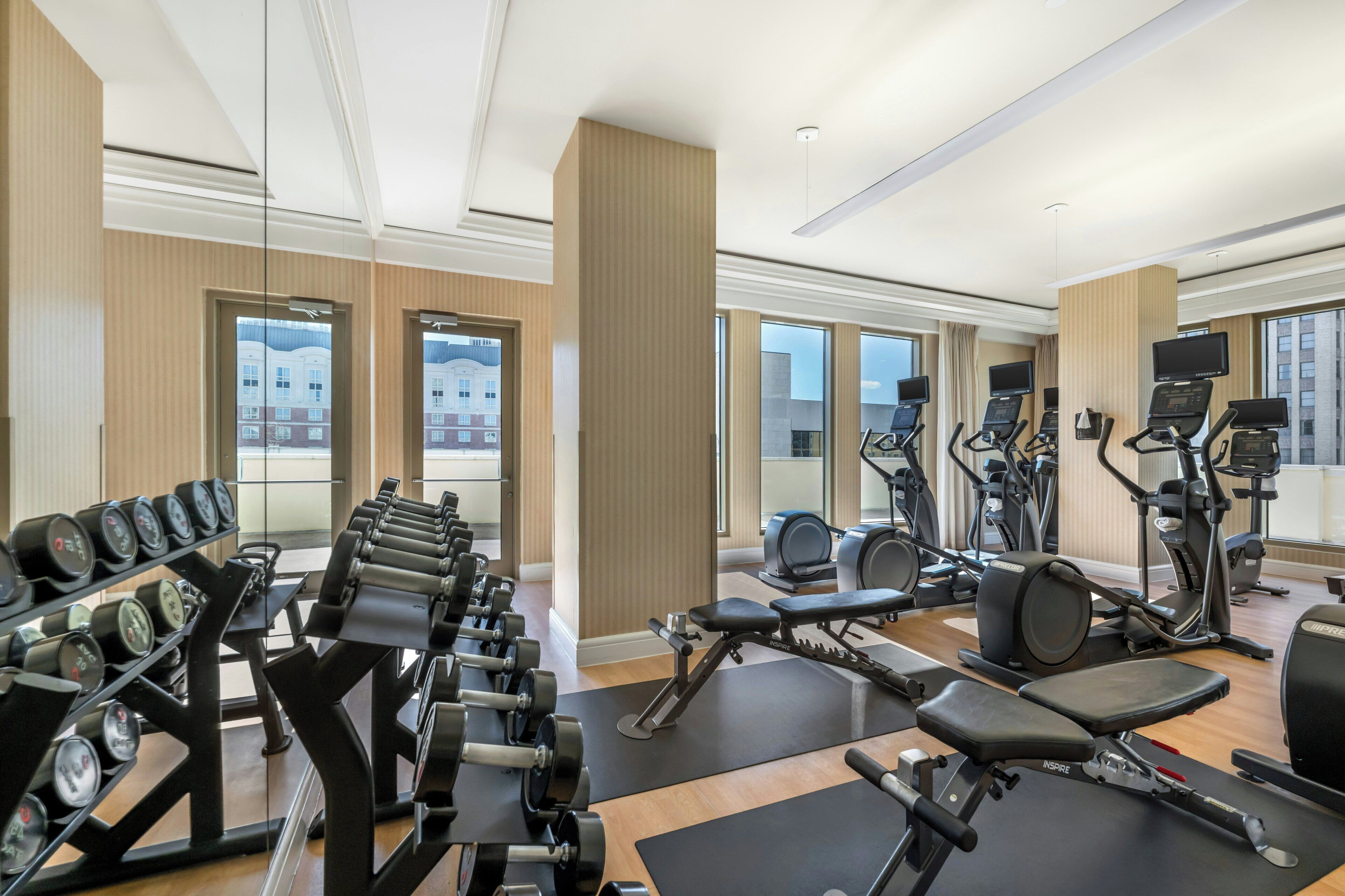 A modern gym with various exercise equipment, large windows, and a view of buildings outside.
