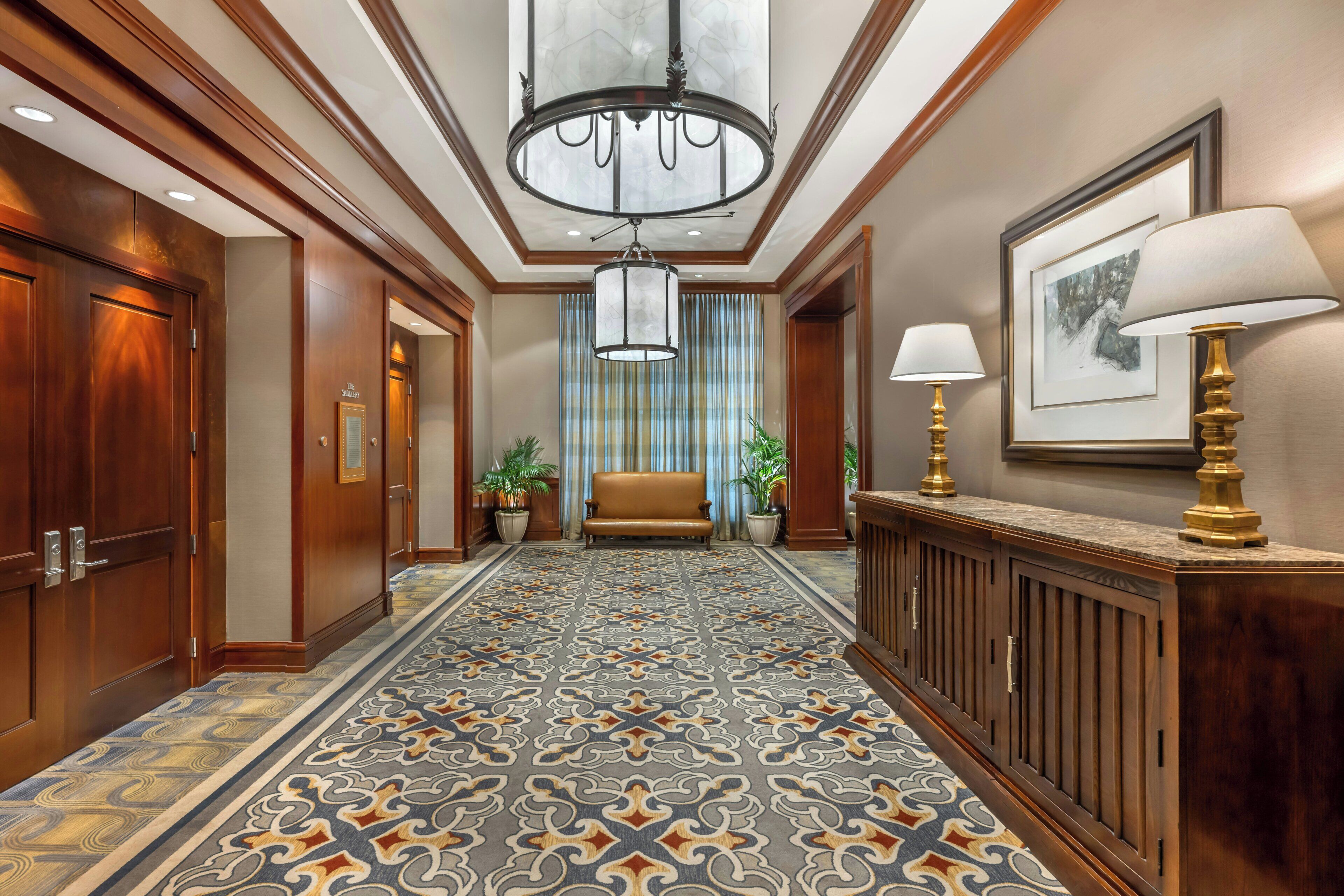 A hotel hallway with a patterned carpet, wooden doors, a sofa, a chandelier, and a lamp.