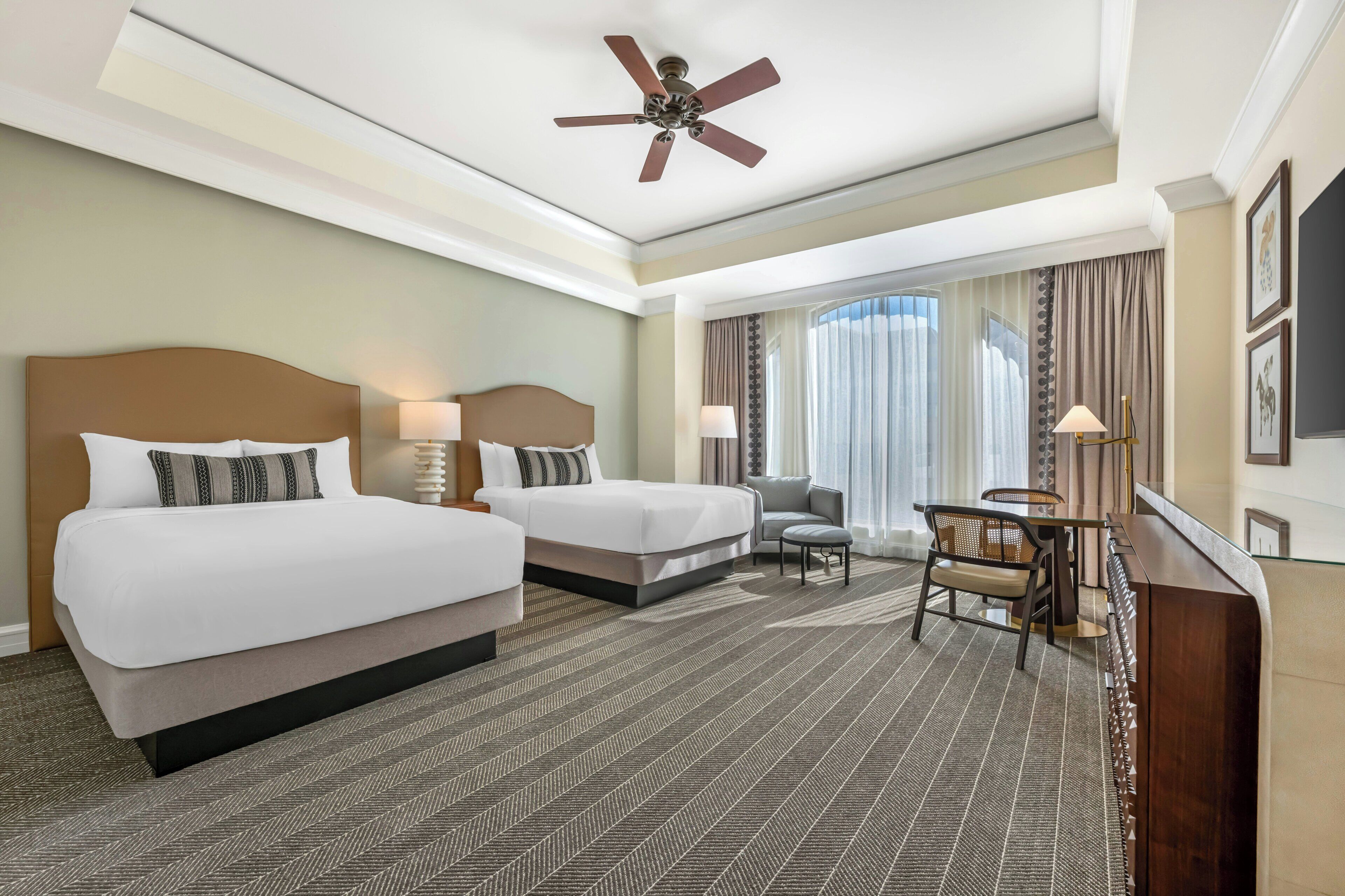 A hotel room with two beds, a desk, a chair, a ceiling fan, and a large window with curtains.