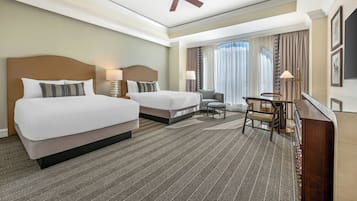 A hotel room with two beds, a desk, a chair, a ceiling fan, and a large window with curtains.