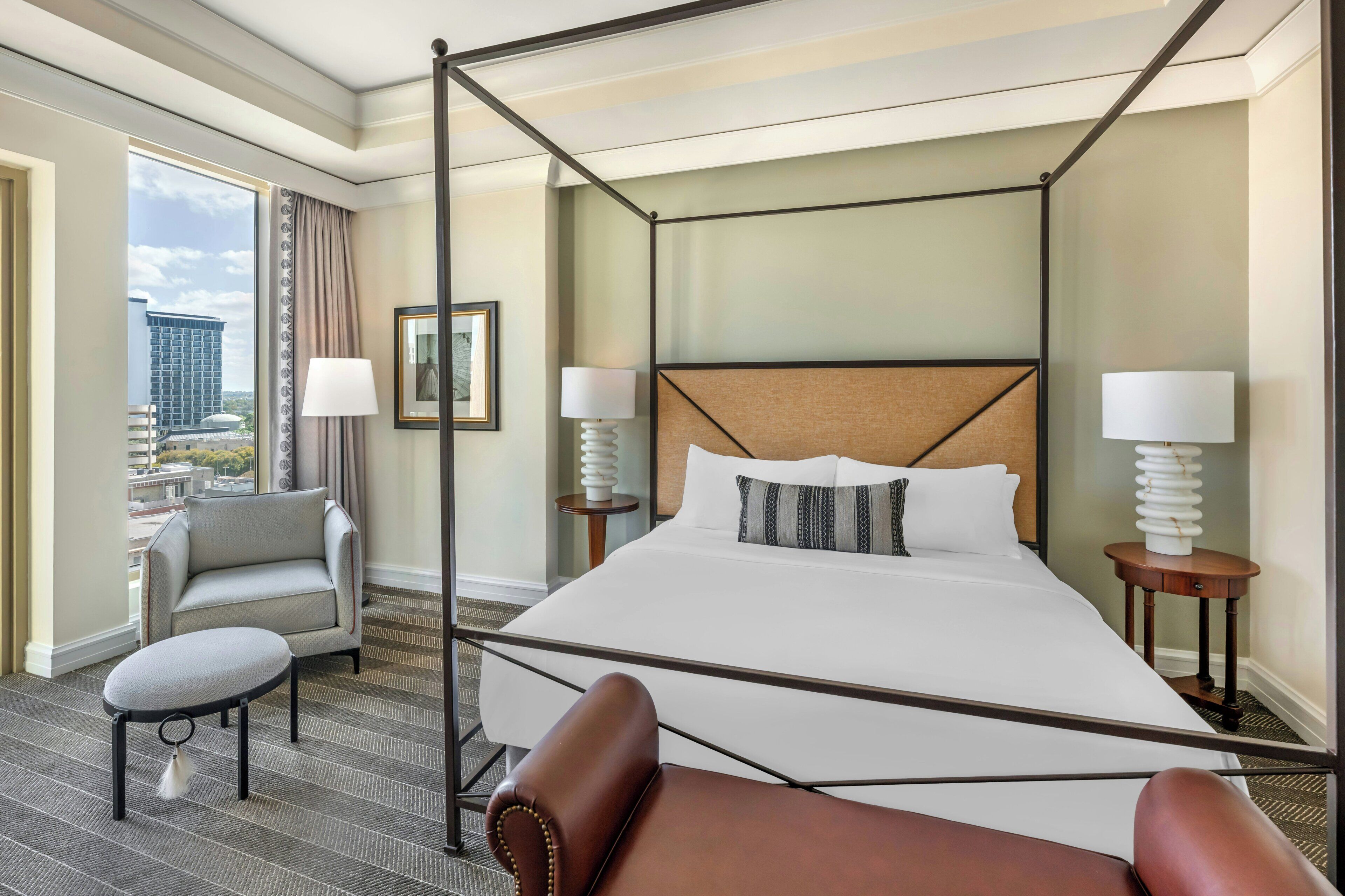 A four-poster bed with a headboard, two bedside tables with lamps, a grey armchair, a small round table, and a view of the city through a large window.