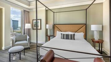 A four-poster bed with a headboard, two bedside tables with lamps, a grey armchair, a small round table, and a view of the city through a large window.