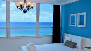 Ein Schlafzimmer mit großem Fenster, einem Bett mit weißer Bettwäsche, einer Chandelierleuchtre und zwei gerahmten Bildern an der Wand.