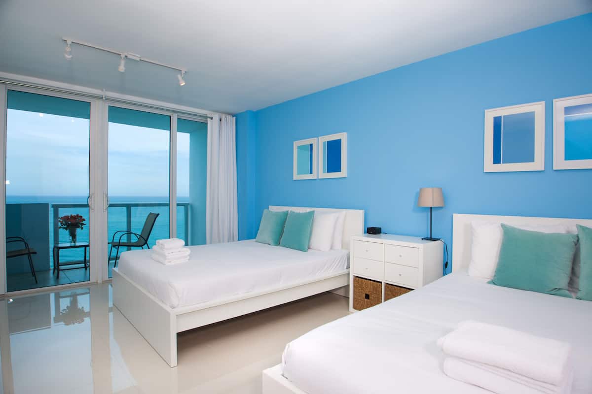 A hotel room with two beds, a balcony with a view, and a white nightstand with a lamp.
