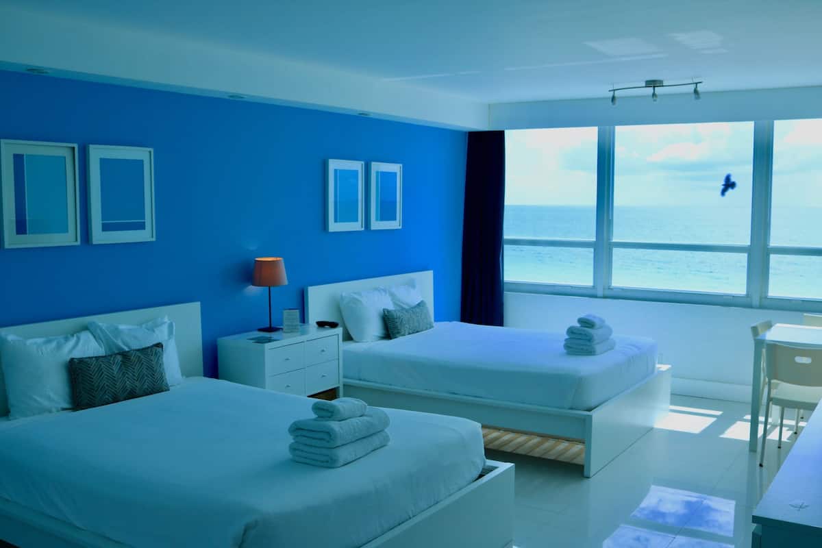 A hotel room with two beds, a desk, and a view of the sea.
