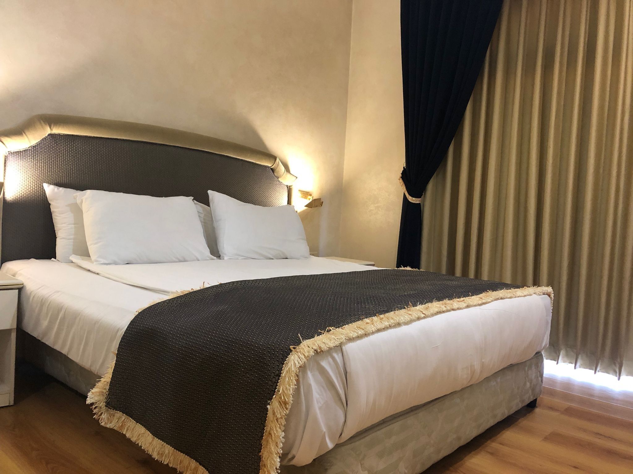 A neatly made bed with white linens and a dark, textured bedspread, a headboard with wall-mounted lighting, and a wooden floor.