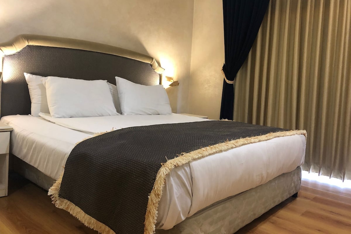 Un lit bien fait, avec du linge de lit blanc et une couvre-lit sombre à motifs, une tête de lit dotée d’un éclairage mural et un plancher en bois.