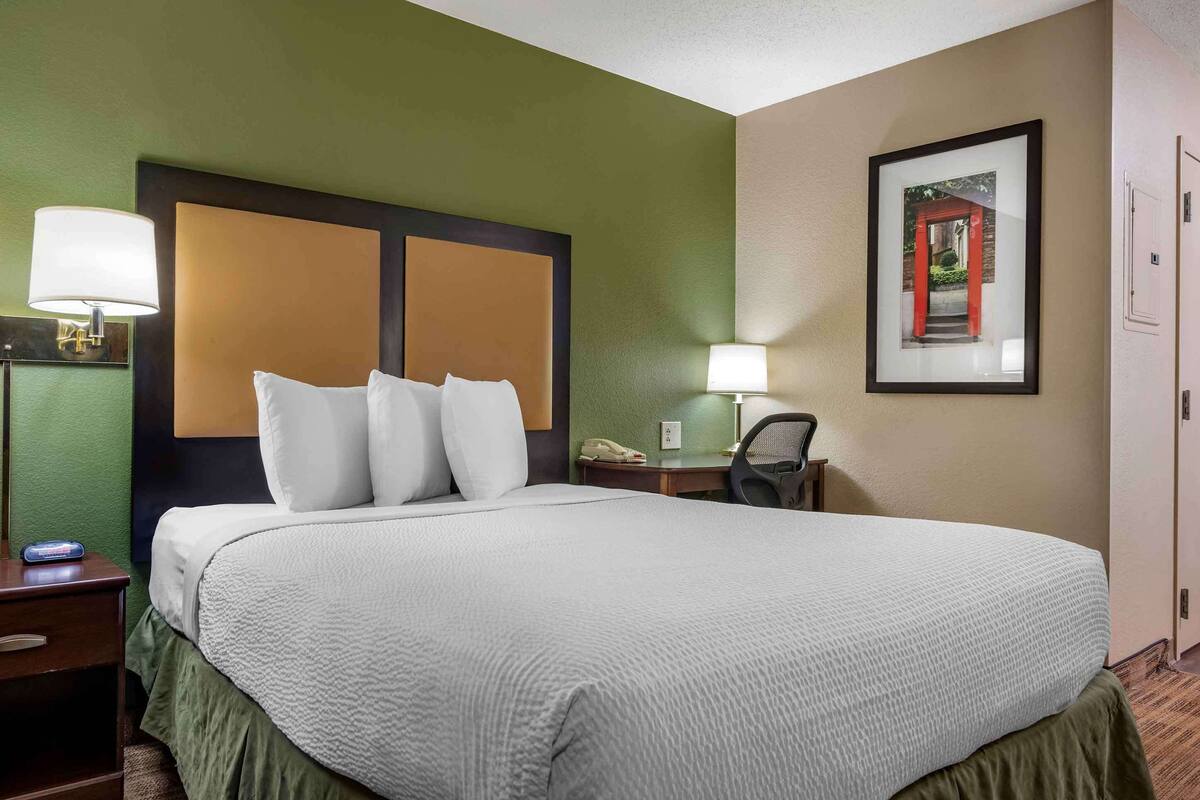 A hotel room with a large bed, two bedside lamps, a desk with a chair, a framed picture on the wall, and a door.