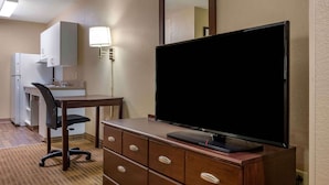 A hotel room with a flat-screen TV on a wooden dresser, a desk with a chair, a kitchenette, and a mirror above the dresser.