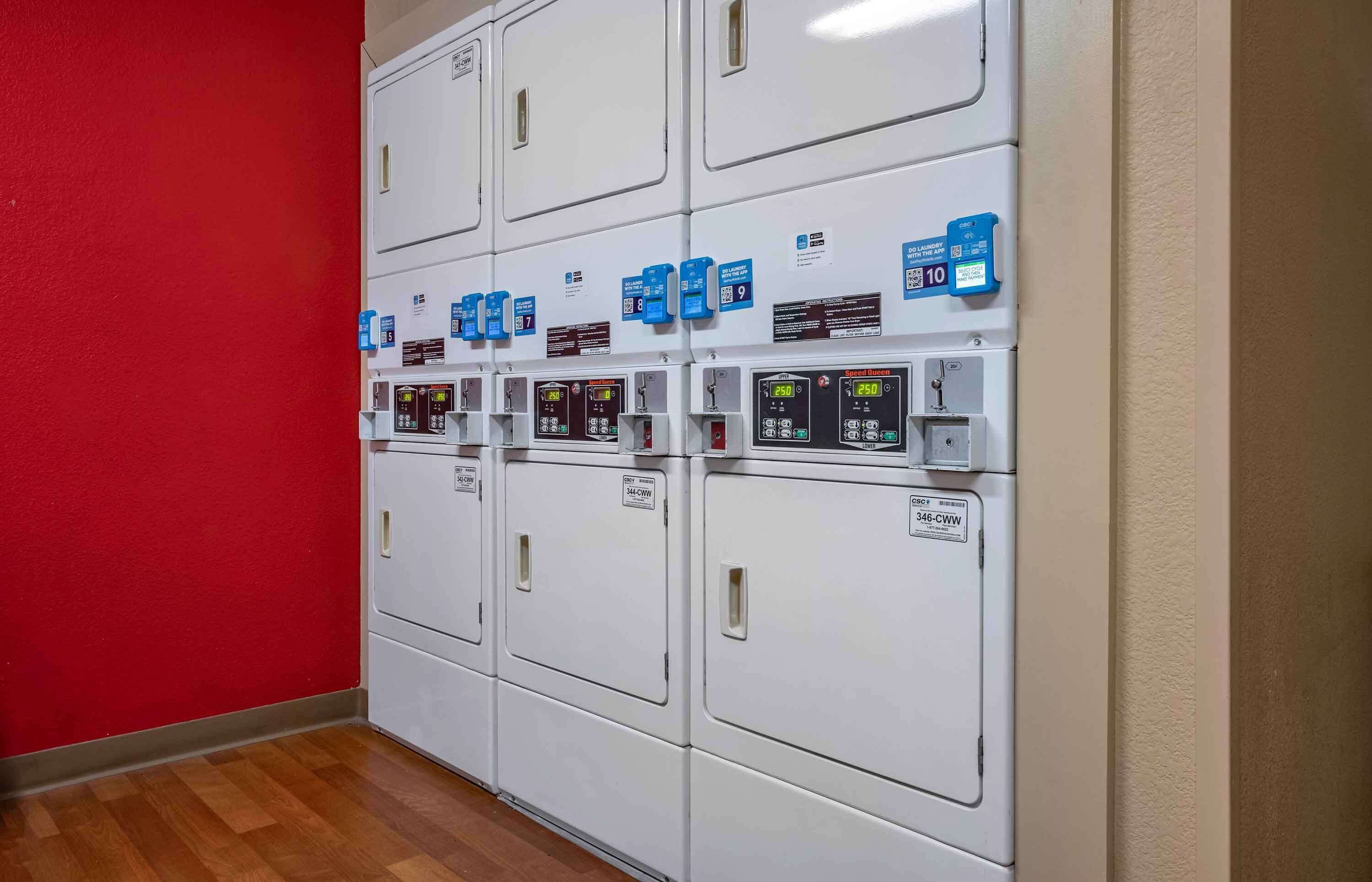 A row of white laundry machines with control panels and blue labels, against a red wall and wooden floor.
