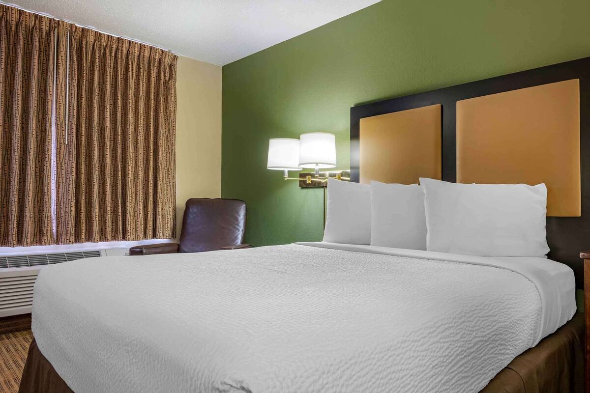 A hotel room with a large bed, a chair, a wall-mounted lamp, and a window with curtains.