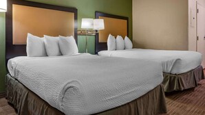 A hotel room with two beds, each with white bedding and a headboard. There is a nightstand with a lamp between the beds.