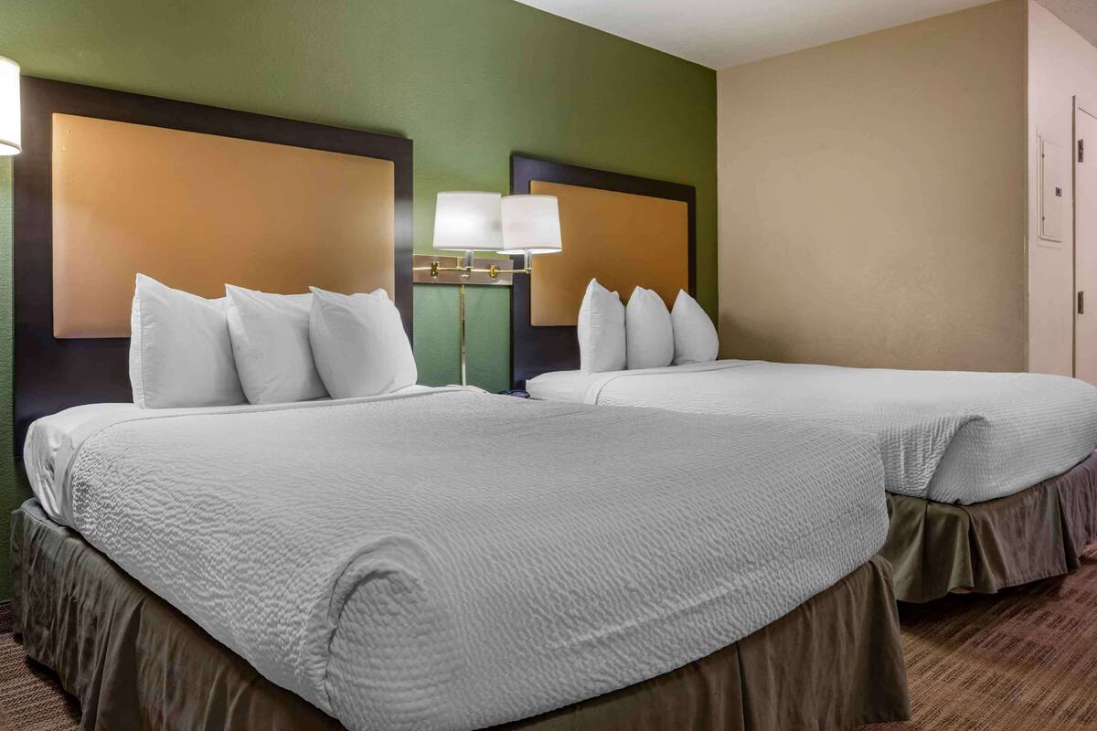 A hotel room with two beds, each with white bedding and a headboard. There is a nightstand with a lamp between the beds.