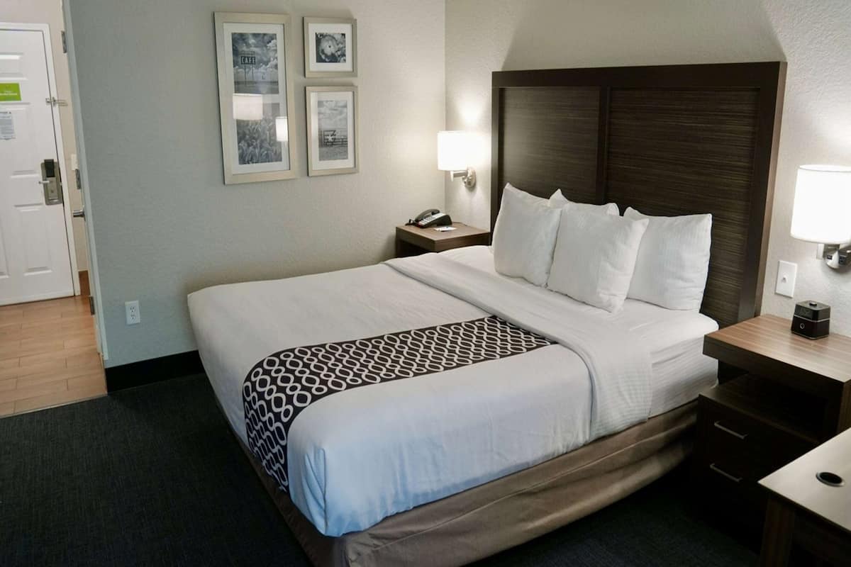 A hotel room with a large bed, a bedside table with a lamp, and a wall-mounted phone. There are framed pictures on the wall and a door to the left.